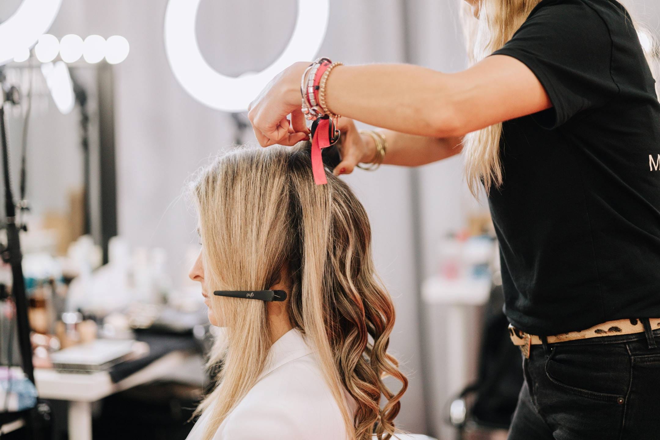 A hairstylist uses a curling iron to style the hair of a seated woman wearing a white jacket and headset in a salon setting.