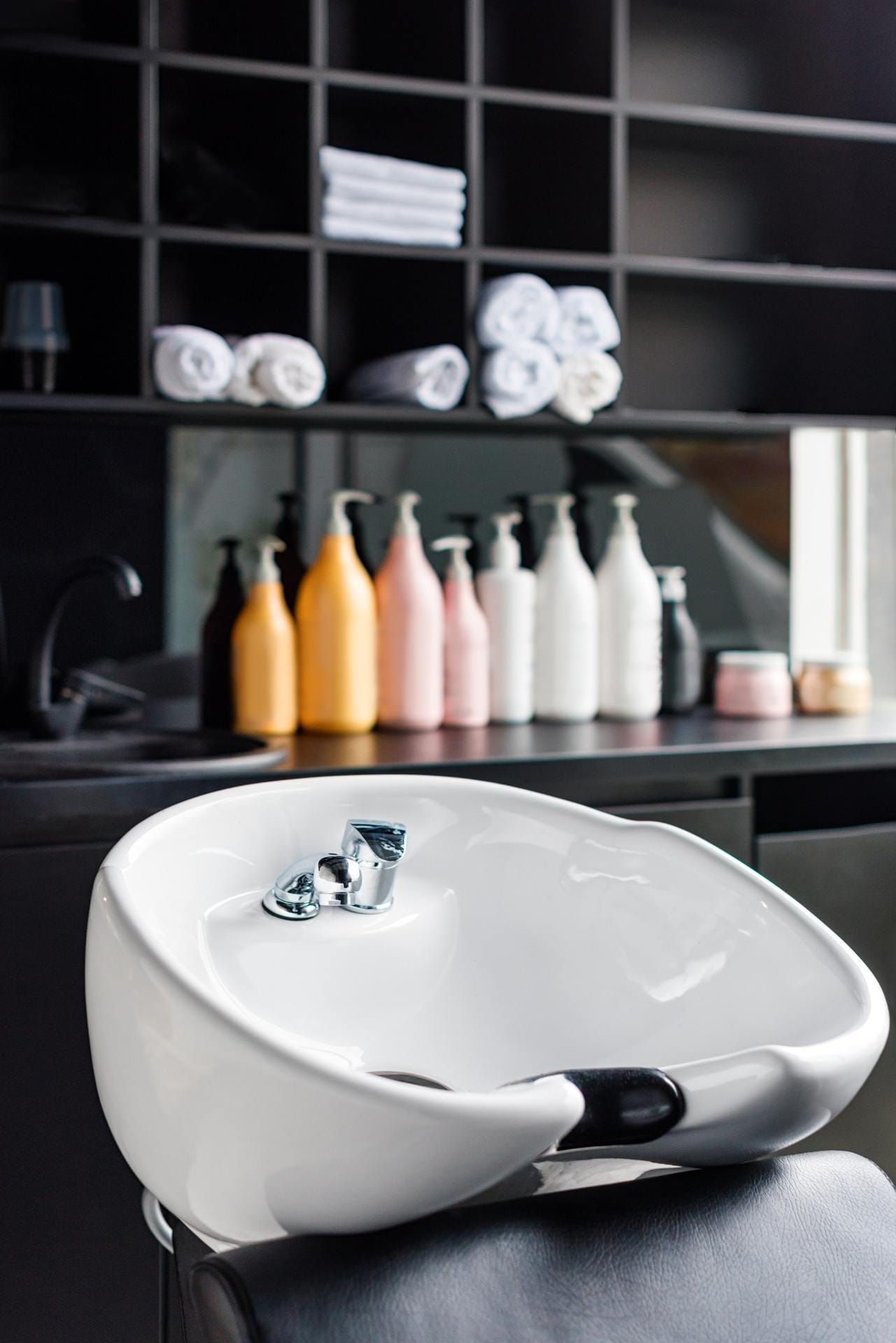 A salon wash basin with a headrest in front of shelves holding rolled towels and bottles of hair products.