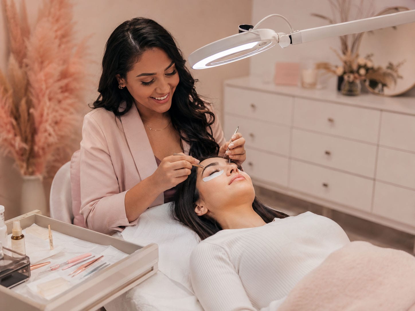 A beauty technician applies eyelash extensions to a client lying on a treatment bed in a well-lit, modern salon featured in the tenant directory.