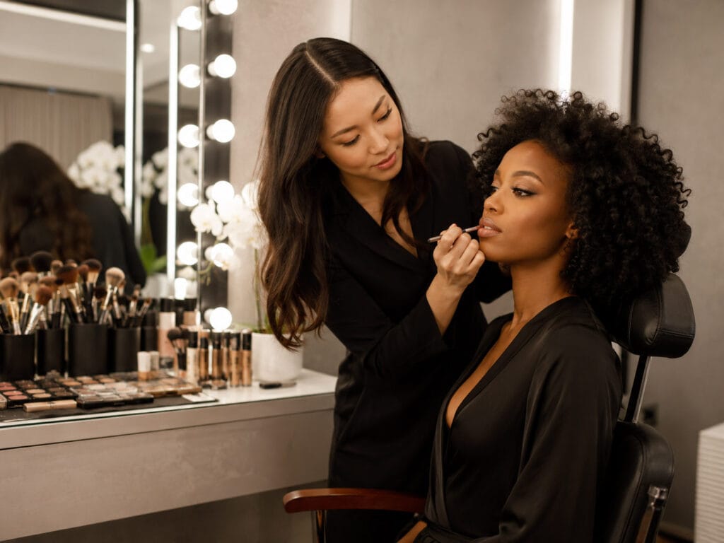 A makeup artist applies lipstick to a seated woman in a dressing room with a lit mirror, makeup products, and brushes on the counter.